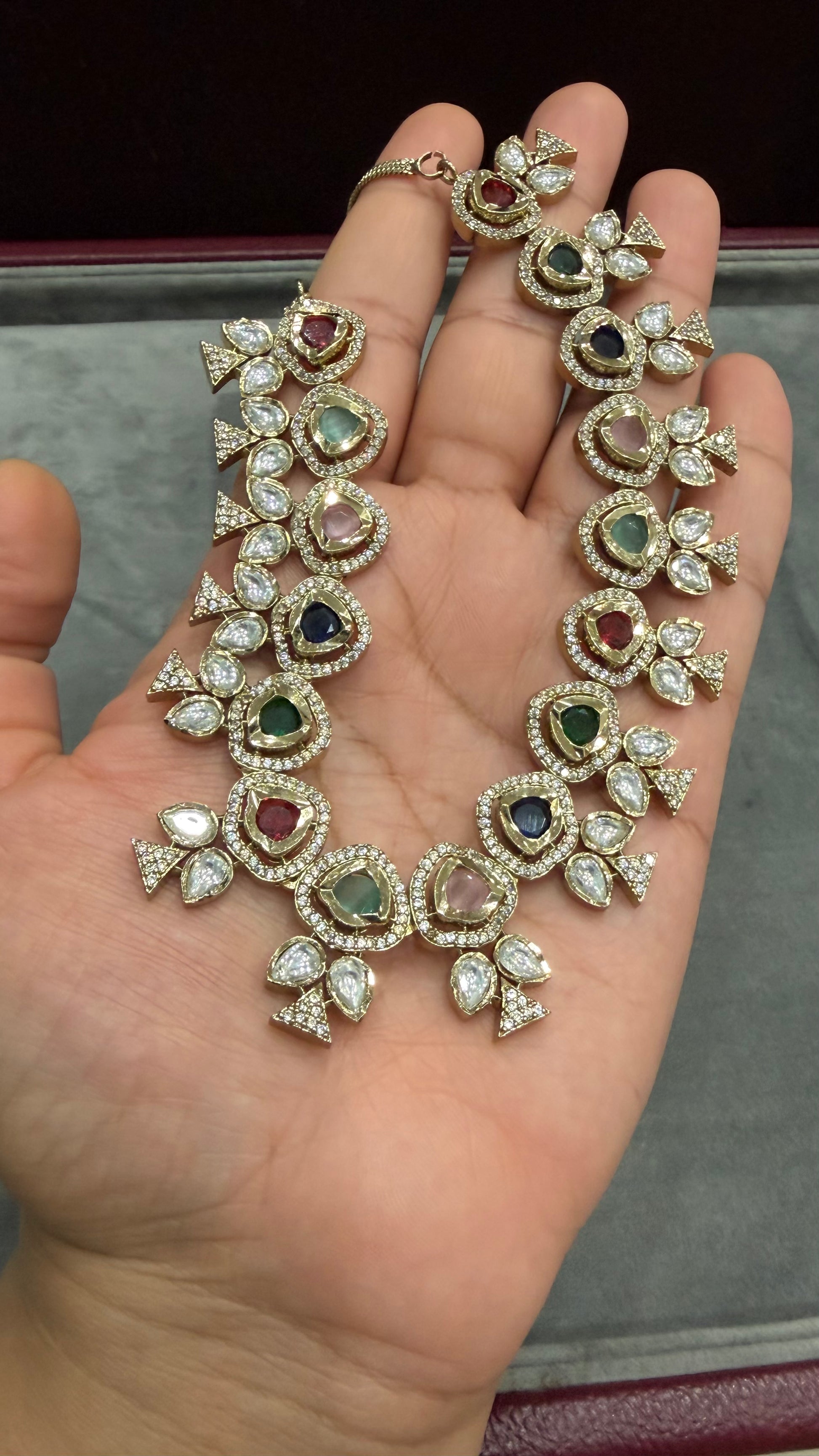 A gold-toned traditional Indian Kundan necklace and matching ring displayed on an open hand. The jewelry features multiple colorful gemstones, including green, pink, blue, and red, set with clear stones in a floral pattern.