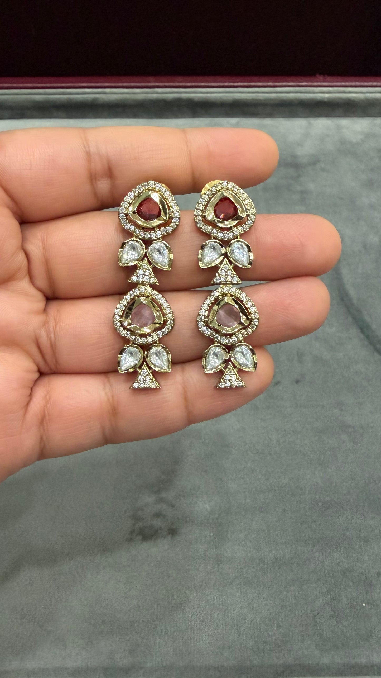 A pair of gold-toned traditional Indian earrings with clear stones and red inlay, held on a person's hand against a gray velvet jewelry box background.