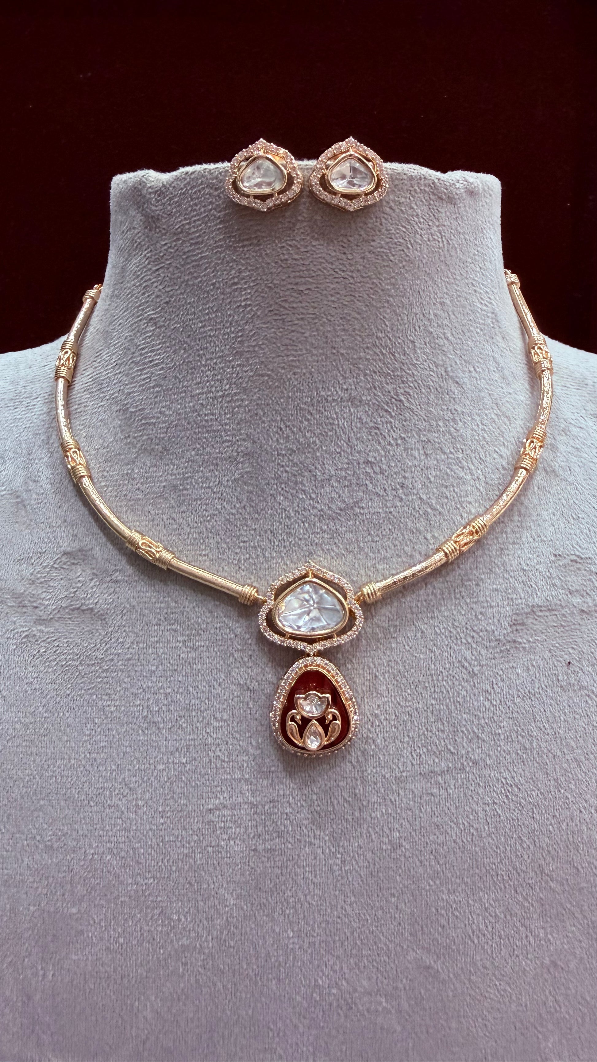 Gold-plated jewelry set featuring a necklace and matching stud earrings displayed on a light gray velvet jewelry bust. The necklace has a beaded chain with a central teardrop pendant featuring a clear heart-shaped stone and a red teardrop accent below it.