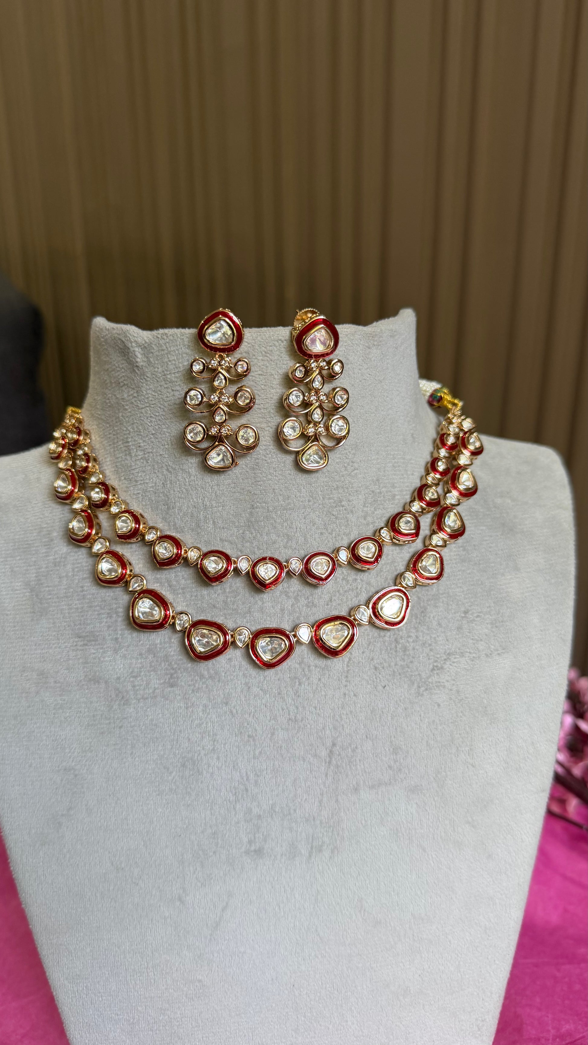 A red and gold kundan jewelry set featuring a double-strand necklace and matching drop earrings, displayed on a light gray velvet jewelry bust.