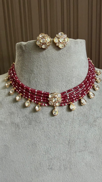 A red beaded necklace and matching stud earrings displayed on a light gray velvet jewelry bust. The necklace features multiple strands of deep red beads, gold-toned metal settings, and dangling clear teardrop stones, with matching flower-shaped stud earrings resting on top of the bust.